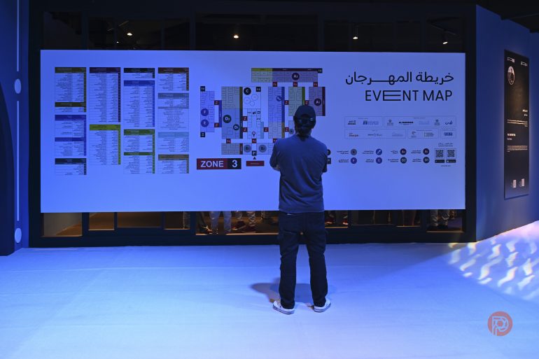 A person stands in front of a large event map displayed on a wall with text in English and Arabic.
