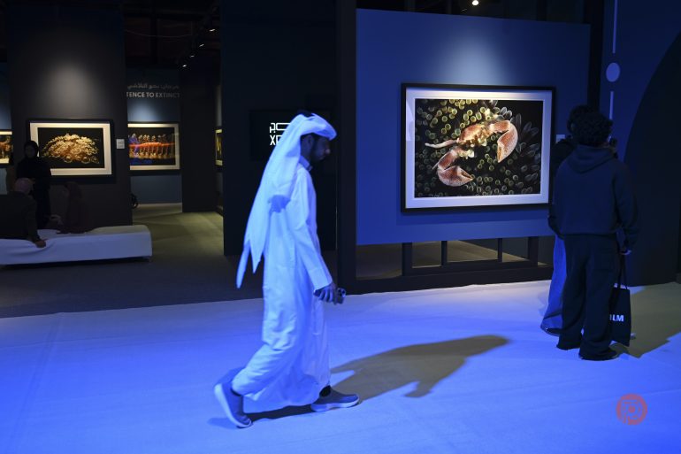 A man in traditional clothing walks past framed artwork at an indoor exhibition with blue lighting.