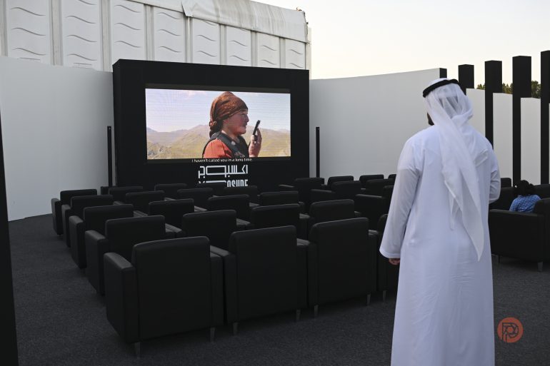 A man in traditional attire stands before empty seats, watching a movie on an outdoor screen.