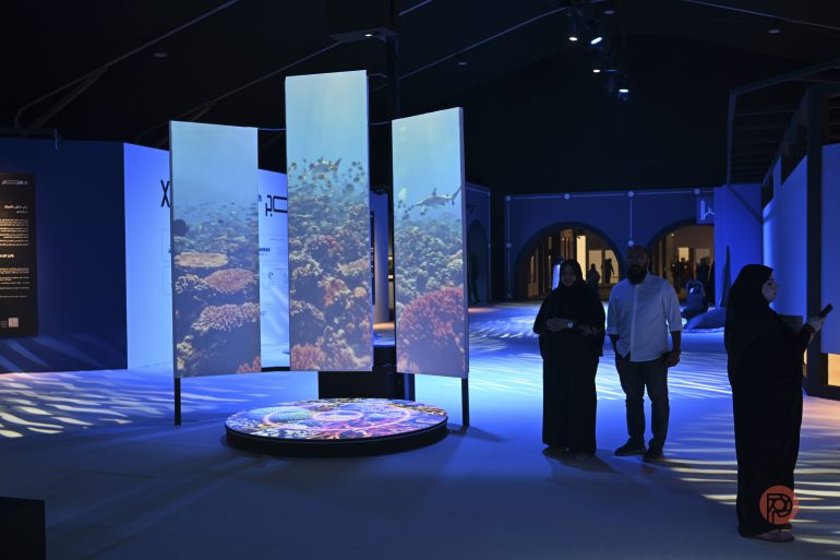 People observing digital panels displaying underwater coral reef scenes in a dimly lit exhibition space.