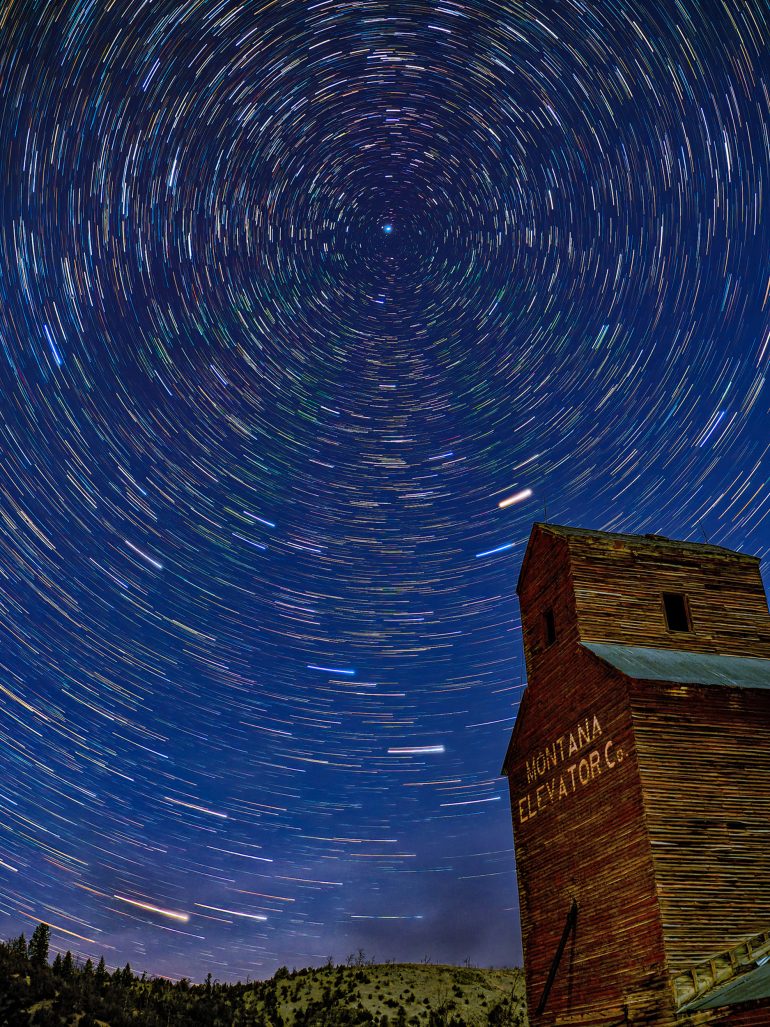 Star trails swirl in the night sky above a rustic wooden Montana Elevator Co. building.