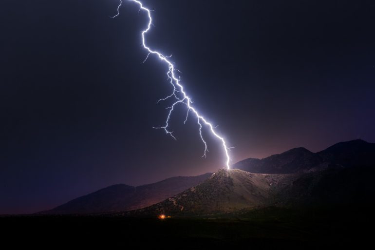 A bolt of lightning strikes the peak of a dark mountain under a night sky.