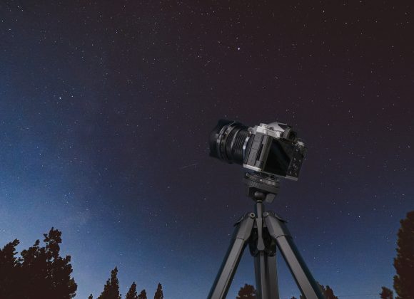 A camera on a tripod pointed at a clear, starry night sky with trees silhouetted below.