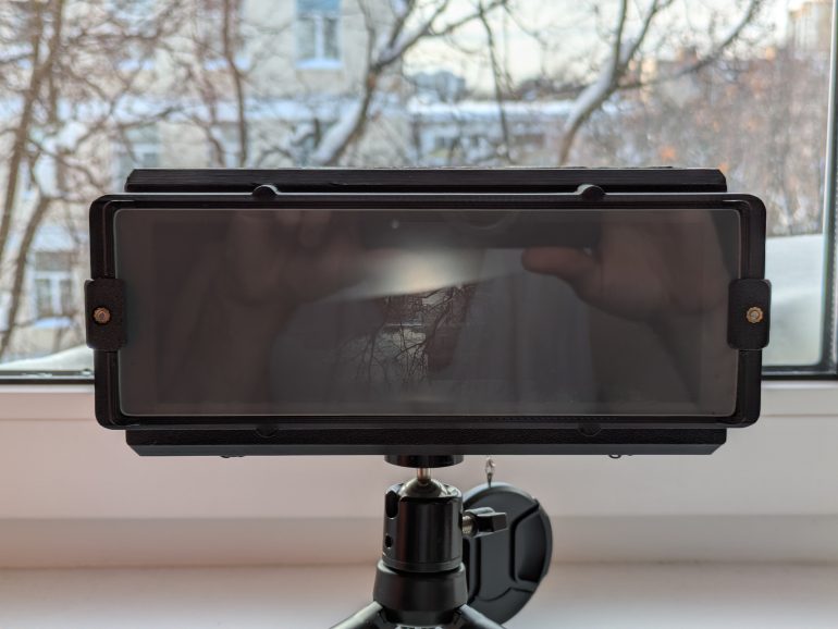 A smartphone mounted horizontally on a tripod near a window with trees outside.
