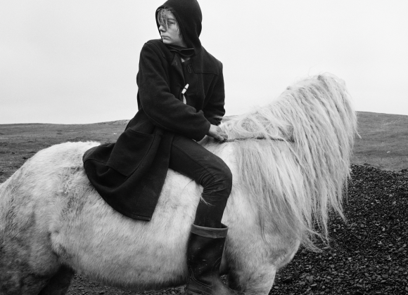 Person in a hooded coat sits sideways on a shaggy white horse, looking into the distance on a barren landscape.