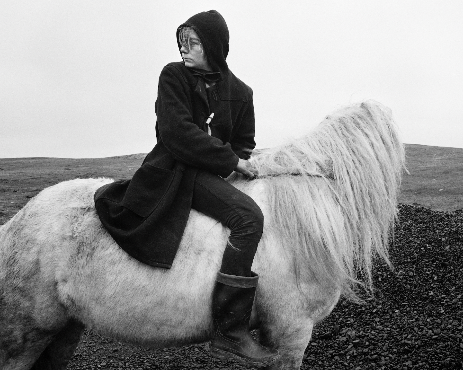 Person in a hooded coat sits sideways on a shaggy white horse, looking into the distance on a barren landscape.