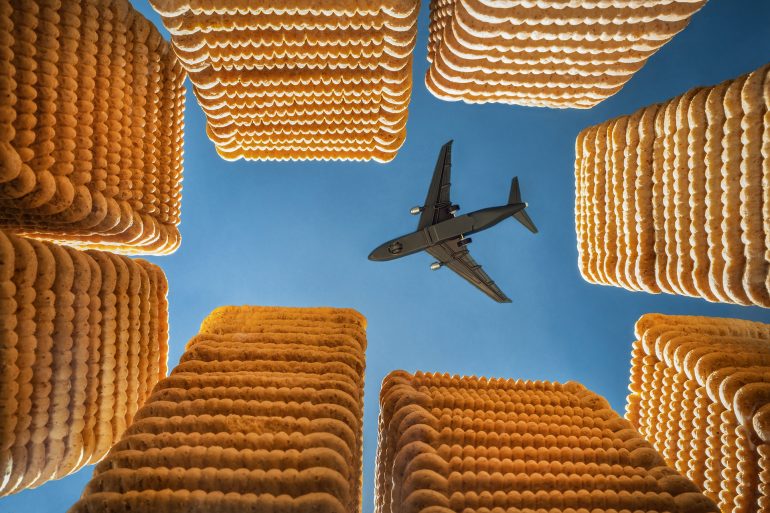 A plane flies overhead, framed by tall stacks of biscuits against a blue sky.
