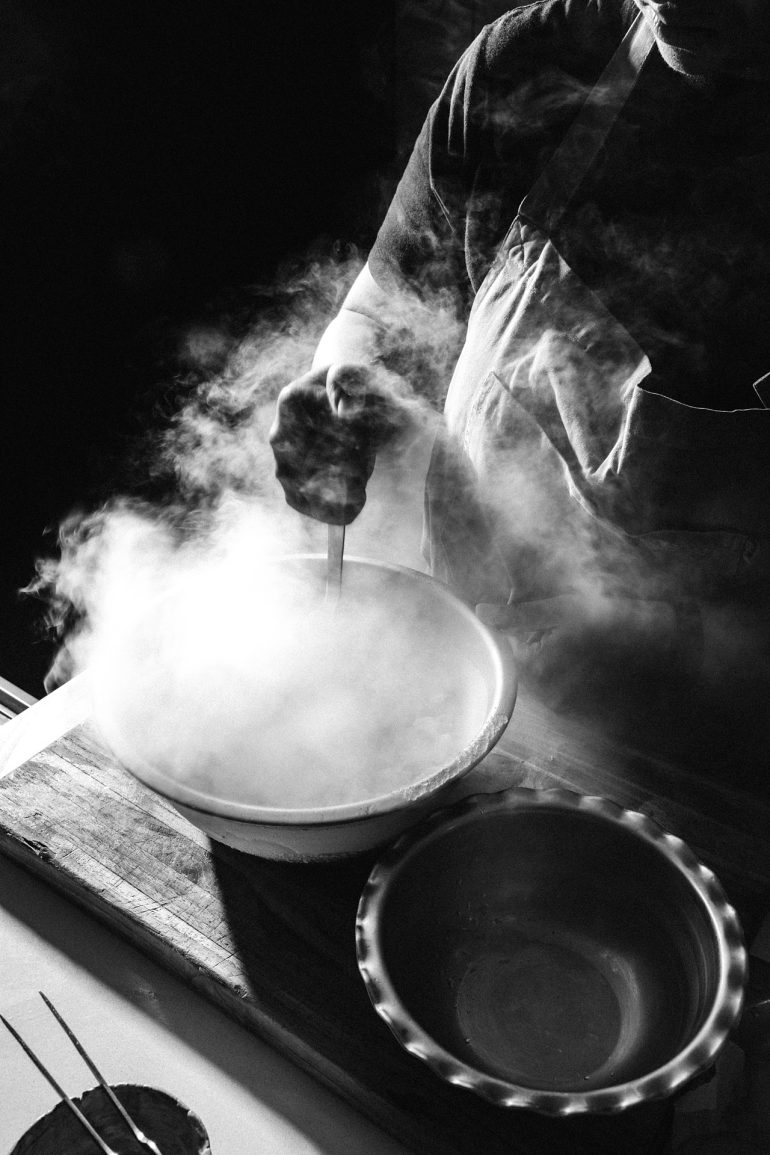 A person stirs a steaming bowl beside an empty metal bowl on a wooden surface, lit dramatically.