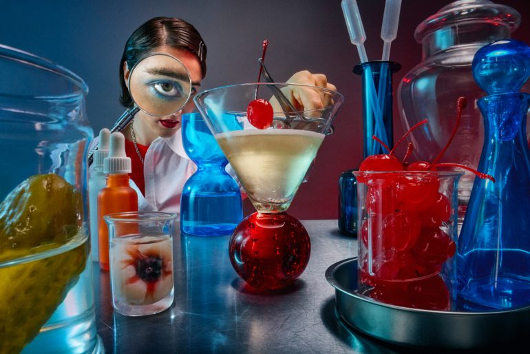 A scientist with a magnified eye adds a cherry to a cocktail amid lab glassware and jars of cherries and pickles.