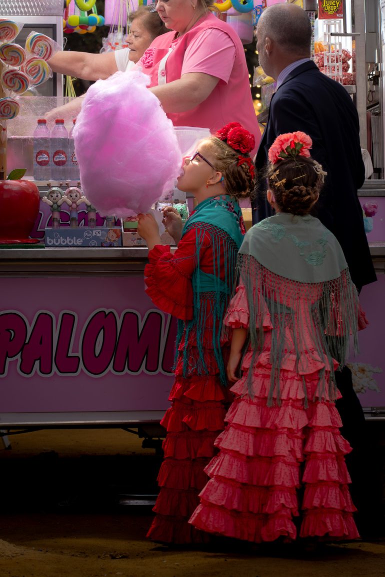 Two girls in ruffled flamenco dresses share cotton candy at a colorful fair booth with adults nearby.