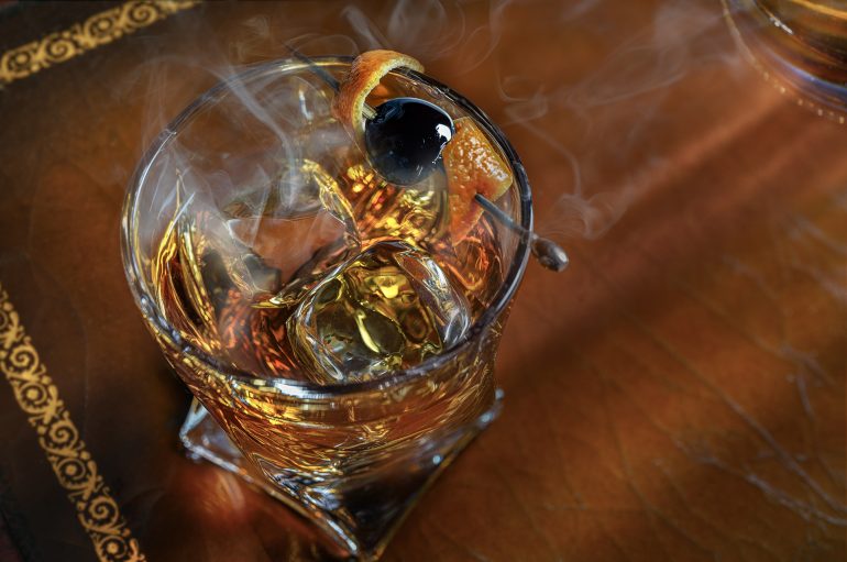 A smoky cocktail with ice, an orange twist, and a cherry garnish in a glass on a brown table.