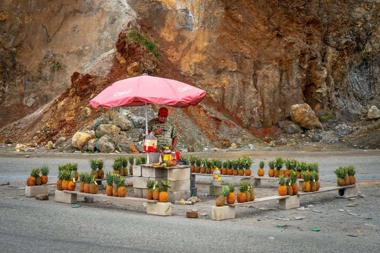 A vendor sells pineapples under a pink umbrella on a roadside, with rocky cliffs in the background.