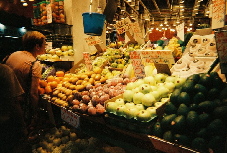 A person browses colorful fruit at a busy market stall with handwritten price signs overhead.