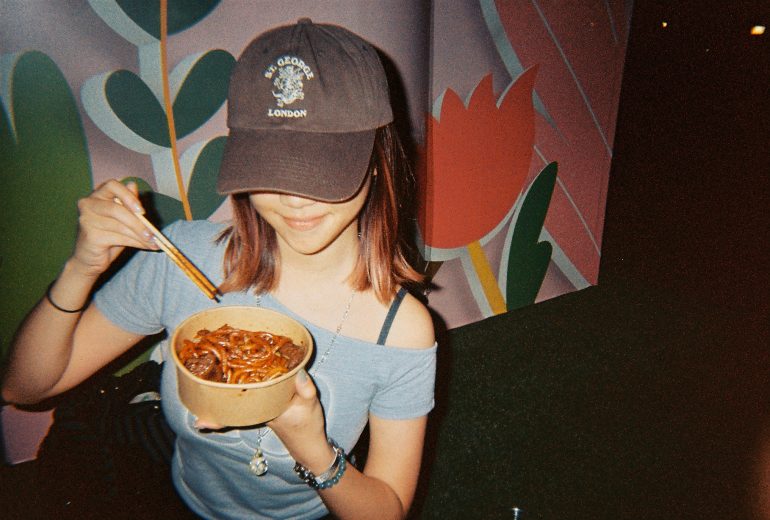 Woman in a cap eating noodles with chopsticks in front of a colorful, floral mural background.
