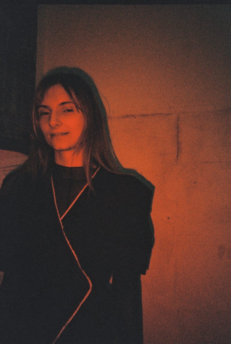 A woman with long hair stands against a red-lit wall, wearing a dark outfit and slight smile.