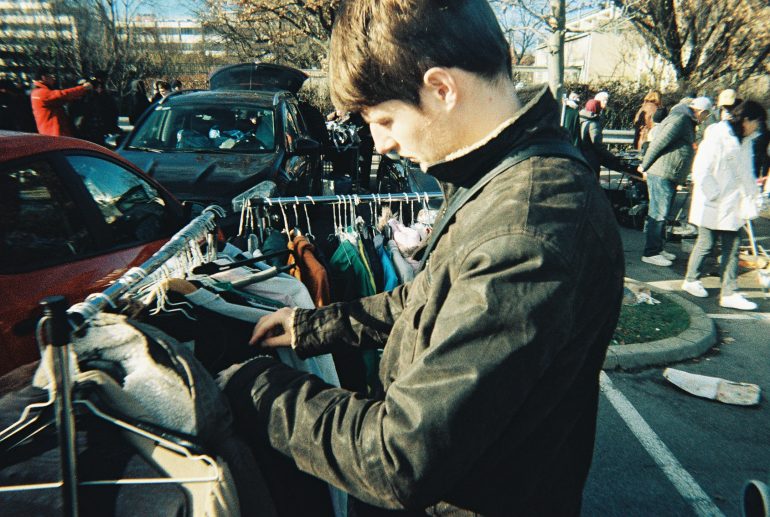 A man browses clothes on a rack at an outdoor market on a sunny day.
