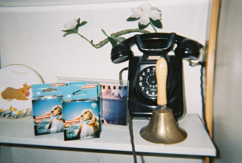 A vintage rotary phone, a brass handbell, and film boxes sit on a white shelf with a flower decoration.