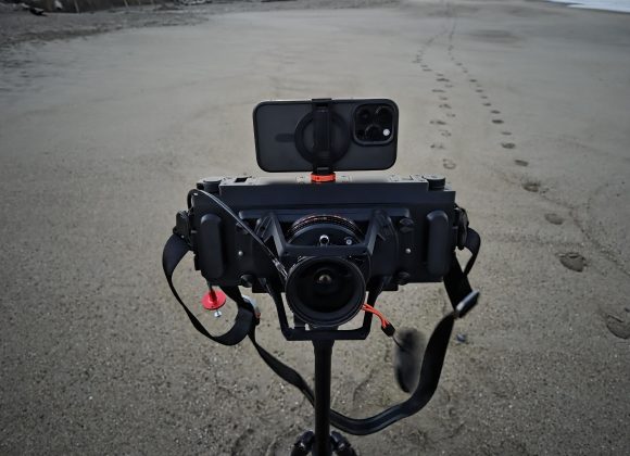 A camera rig with a smartphone mounted on top, set up on a tripod on a sandy beach.