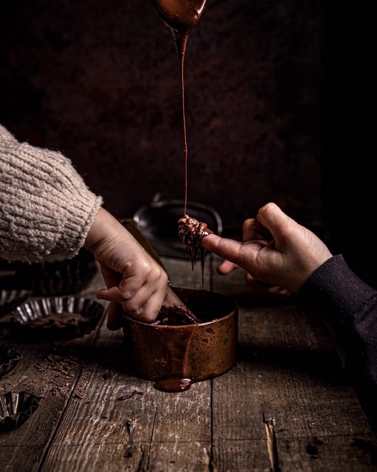 Two hands dip into a pot of melted chocolate, one licking chocolate off their finger.