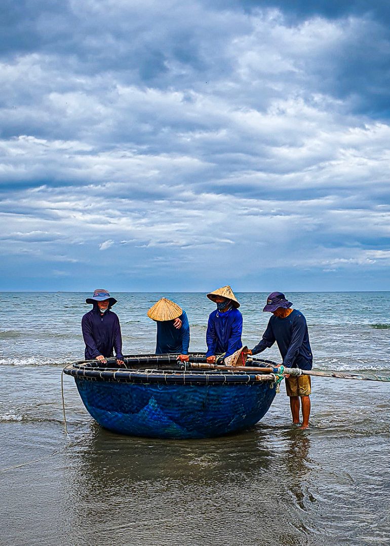 Four people in hats stand by a round blue boat on the shore, with cloudy sky and calm sea in the background.