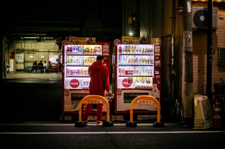 A person in red stands at two brightly lit vending machines at night on a dimly lit street.