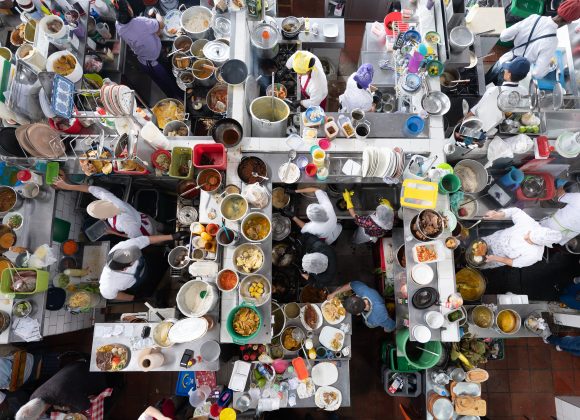 A busy overhead view of chefs preparing food at crowded kitchen stations filled with dishes and ingredients.