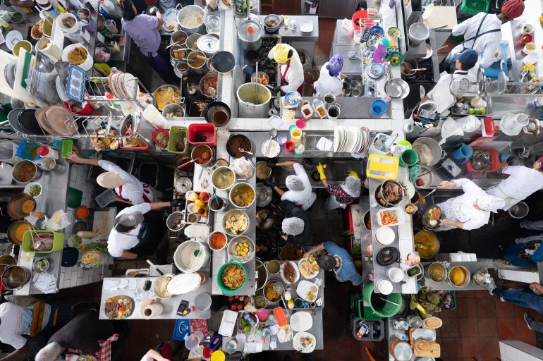 A busy overhead view of chefs preparing food at crowded kitchen stations filled with dishes and ingredients.