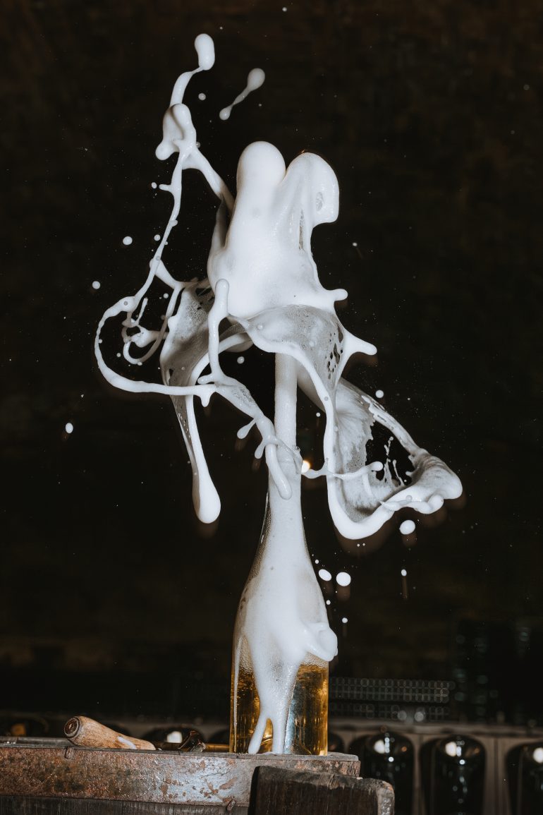 Foamy beer erupts dramatically from a bottle, splashing white foam in mid-air against a dark background.