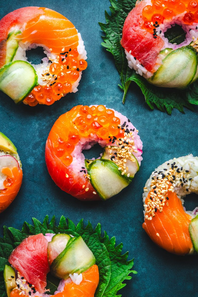Colorful sushi donuts with salmon, tuna, cucumber, roe, and sesame seeds on green leaves over a blue surface.