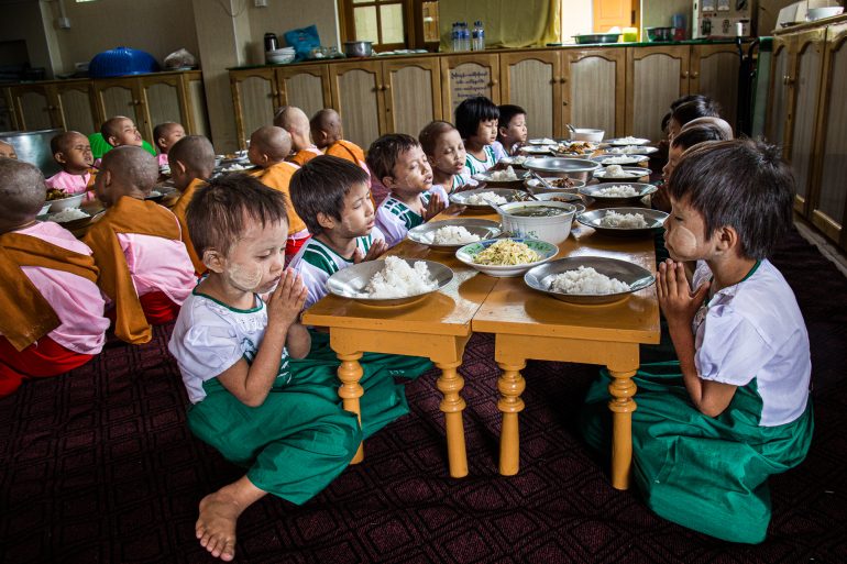 Young children with shaved heads sit on the floor, praying before meals at low tables in a dining hall.