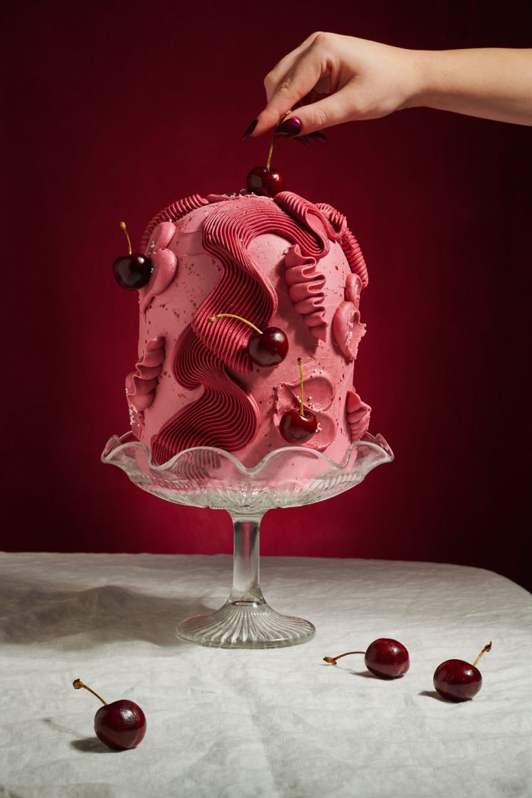 A hand places a cherry on a tall pink cake with swirled icing, on a glass stand, against a red background.