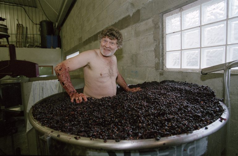 A shirtless man stands in a large vat filled with grapes inside a winery or industrial building.