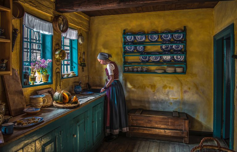 A woman in traditional dress works in a rustic kitchen with yellow walls and blue cabinets, near a window.
