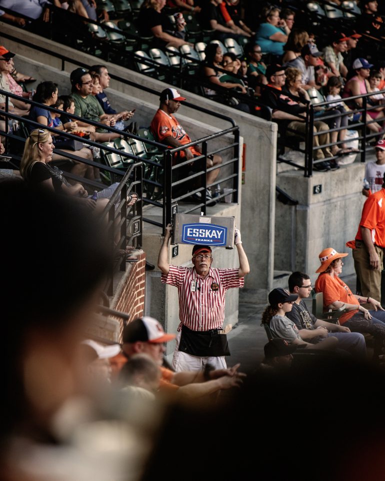 A vendor in a striped shirt holds an ESSKAY box above his head at a crowded baseball stadium.