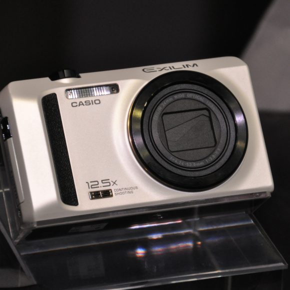 A silver Casio Exilim digital camera with a 12.5x zoom lens displayed on a clear stand.