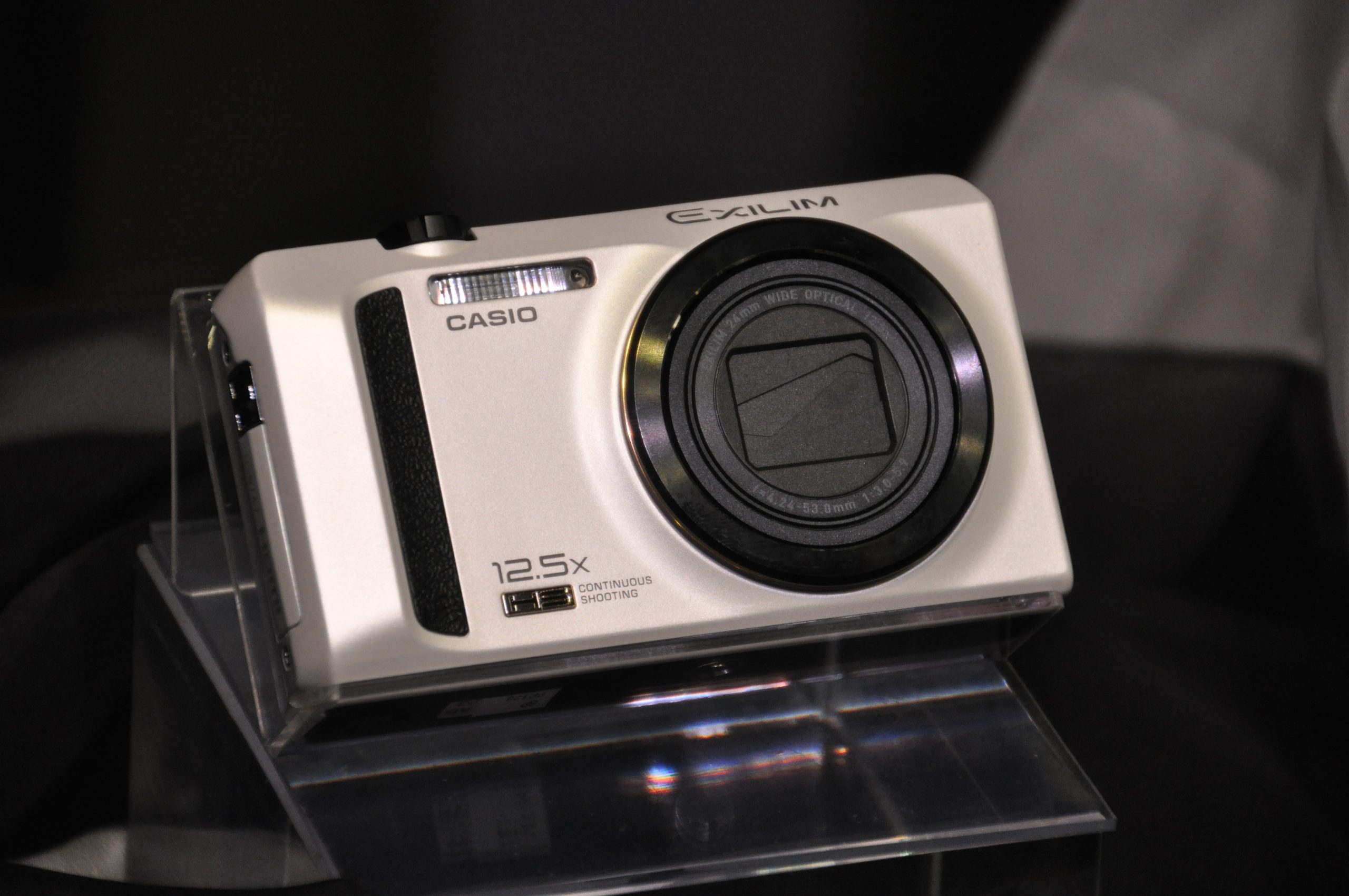 A silver Casio Exilim digital camera with a 12.5x zoom lens displayed on a clear stand.