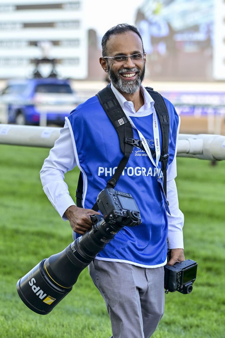 A smiling photographer wearing a blue vest stands on grass holding a camera with a large lens.