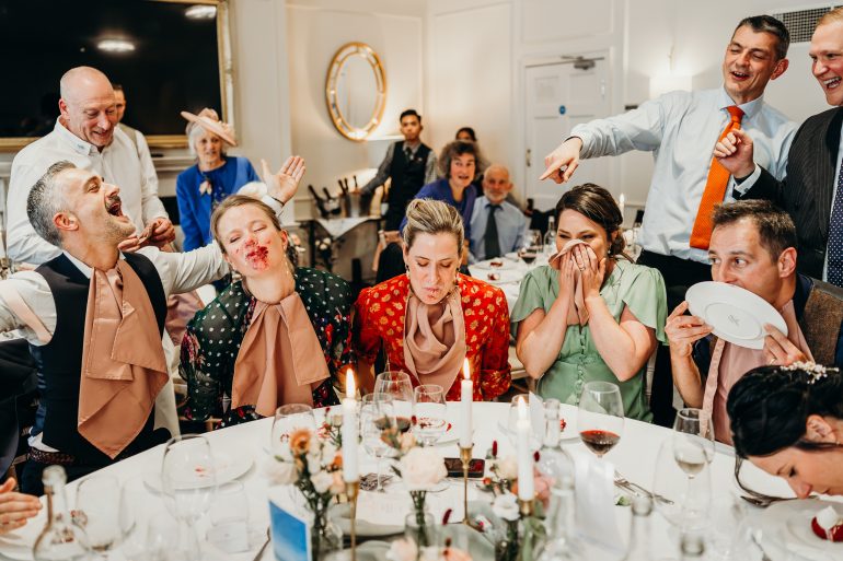 A group at a lively party sits around a table, laughing as three people have cake on their faces.