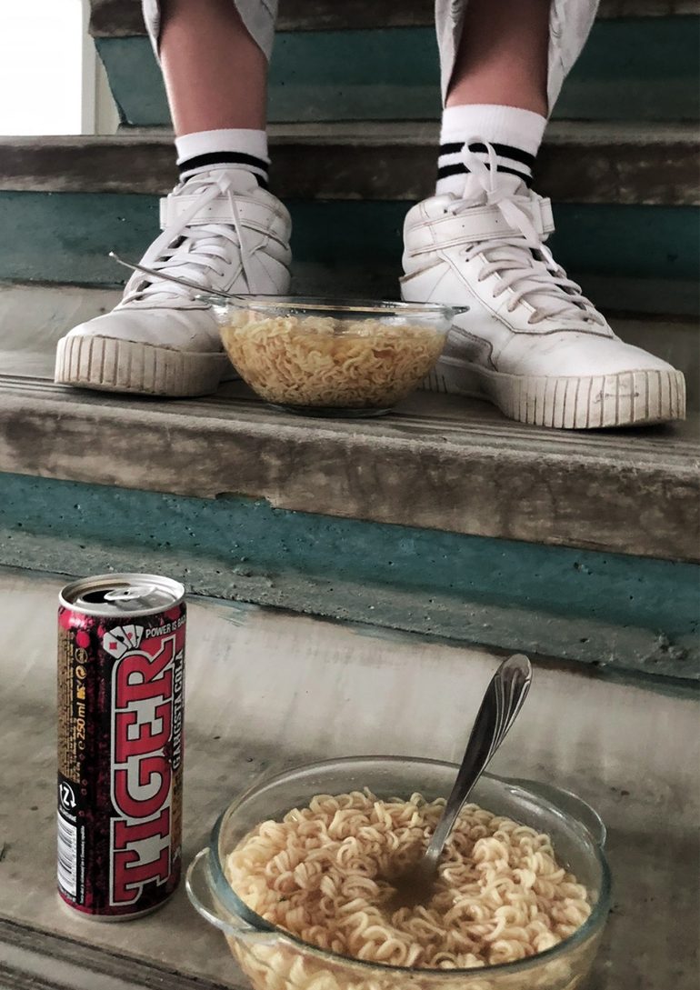 Two bowls of instant noodles and a can of Tiger energy drink on stairs, with a person wearing white sneakers.