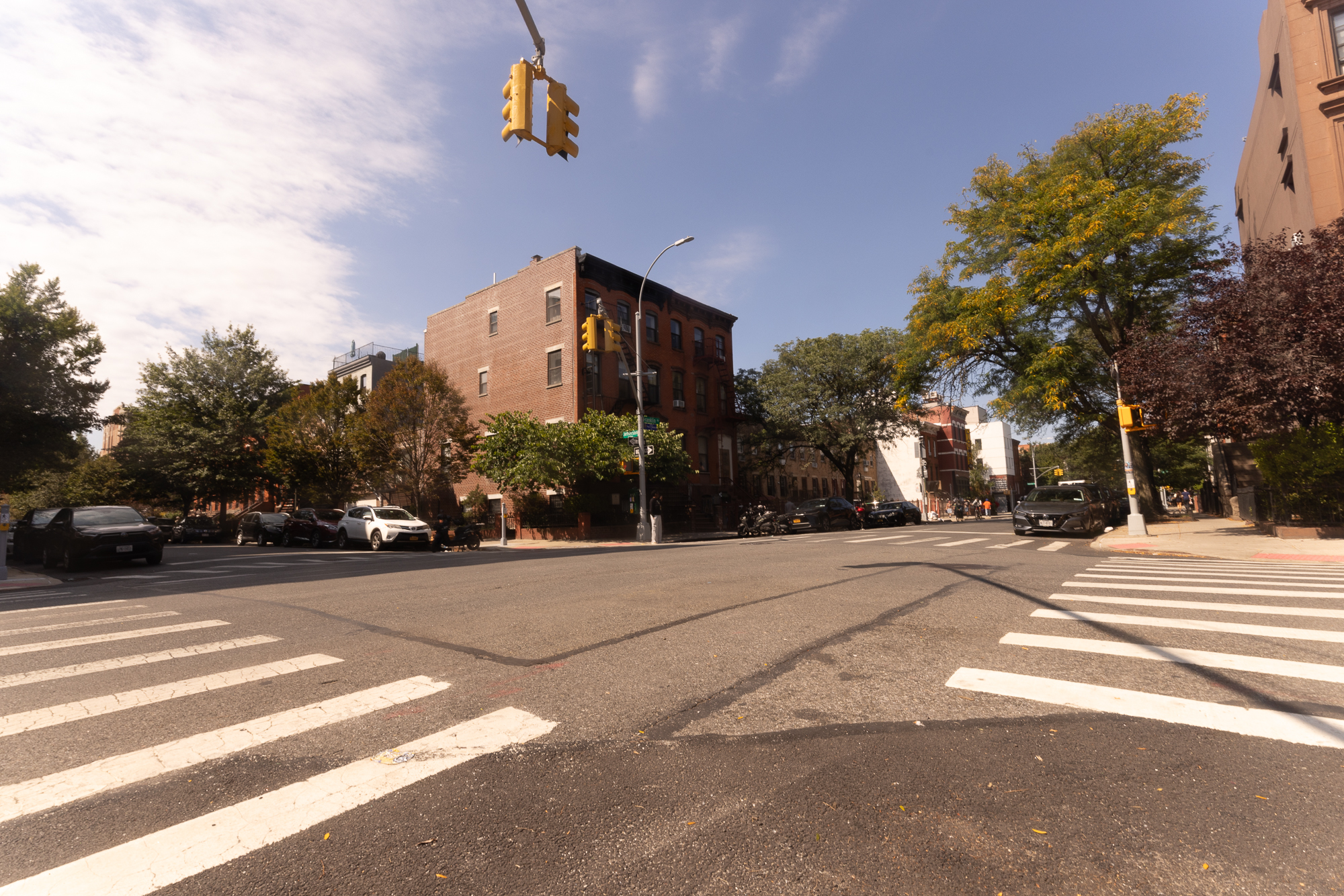 A sunny urban street intersection with crosswalks, parked cars, trees, and brick apartment buildings.