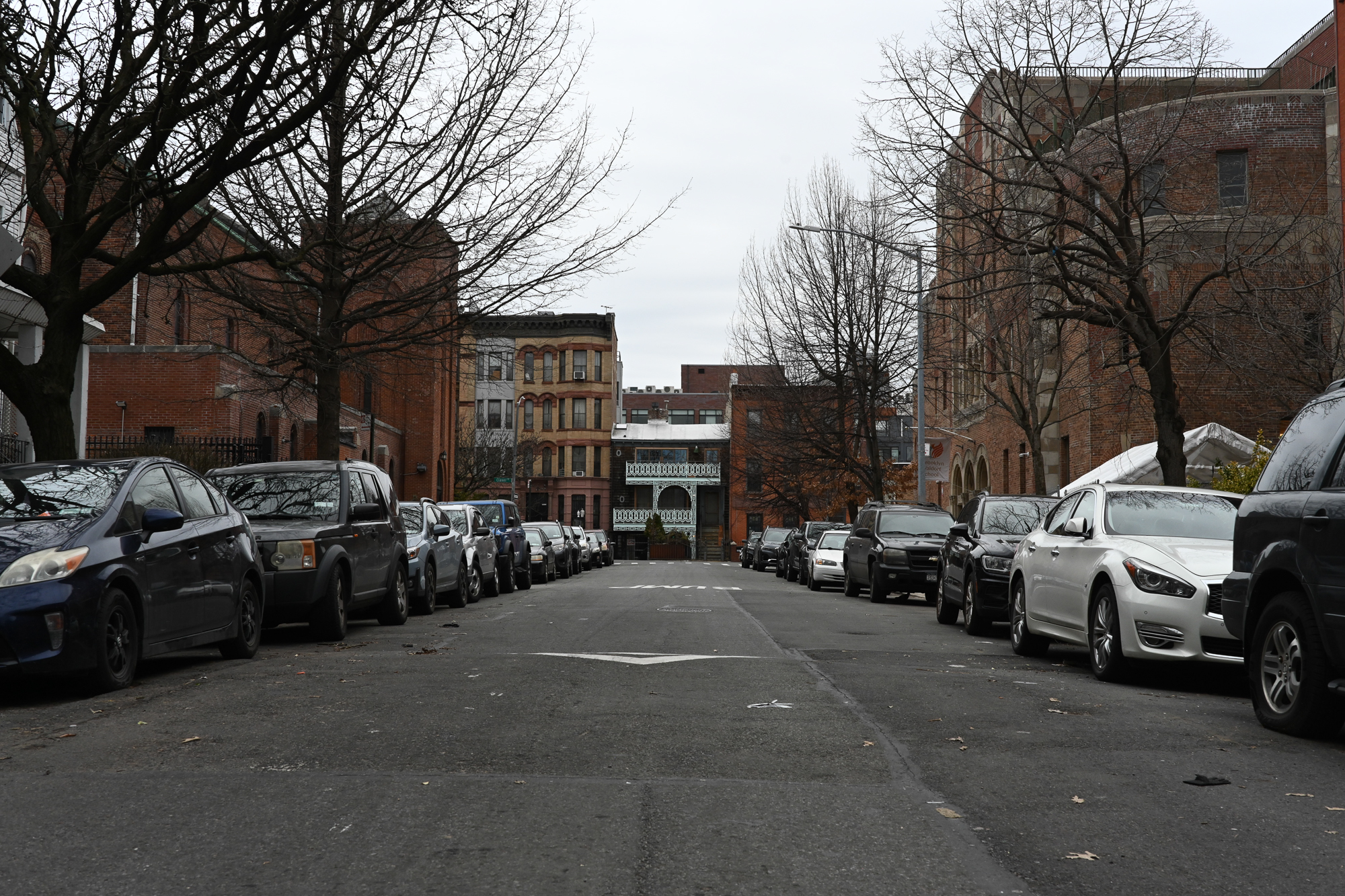 A quiet city street lined with parked cars, bare trees, and brick buildings on a cloudy day.