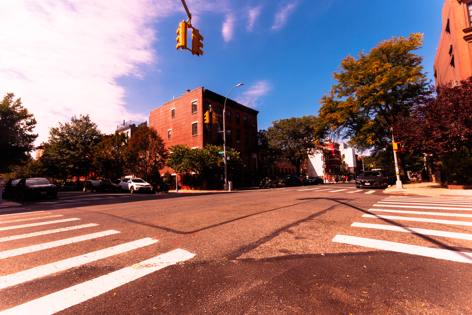 A sunny urban street intersection with crosswalks, traffic lights, and brick buildings under a blue sky.