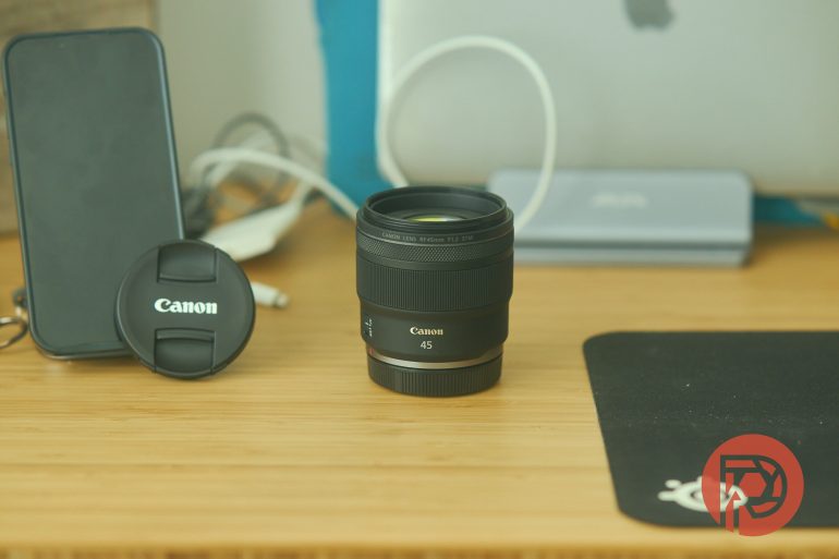A Canon 45mm camera lens on a desk beside its lens cap, a phone, and a mouse pad.