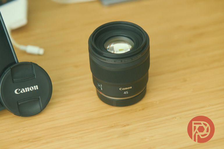 A Canon camera lens and lens cap on a wooden surface with a red logo in the corner.