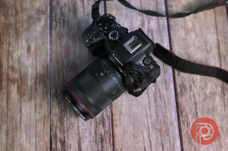A Canon camera with a zoom lens rests on a wooden surface, viewed from above.