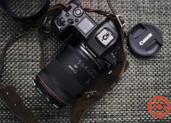Canon camera with attached lens and leather strap on woven surface, Canon lens cap nearby, red logo in corner.