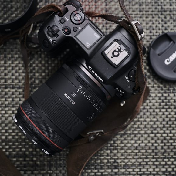 Canon camera with attached lens and leather strap on woven surface, Canon lens cap nearby, red logo in corner.