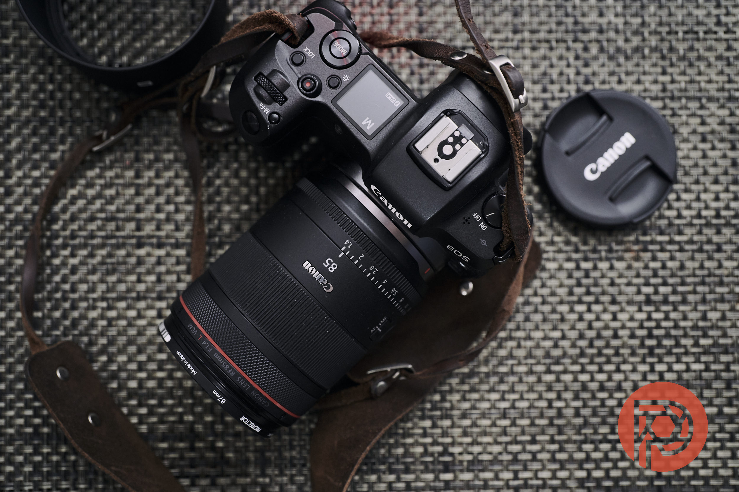Canon 85mm F1.4 L VCM Review: Why Does Canon Hate Their Customers? Canon camera with attached lens and leather strap on woven surface, Canon lens cap nearby, red logo in corner.
