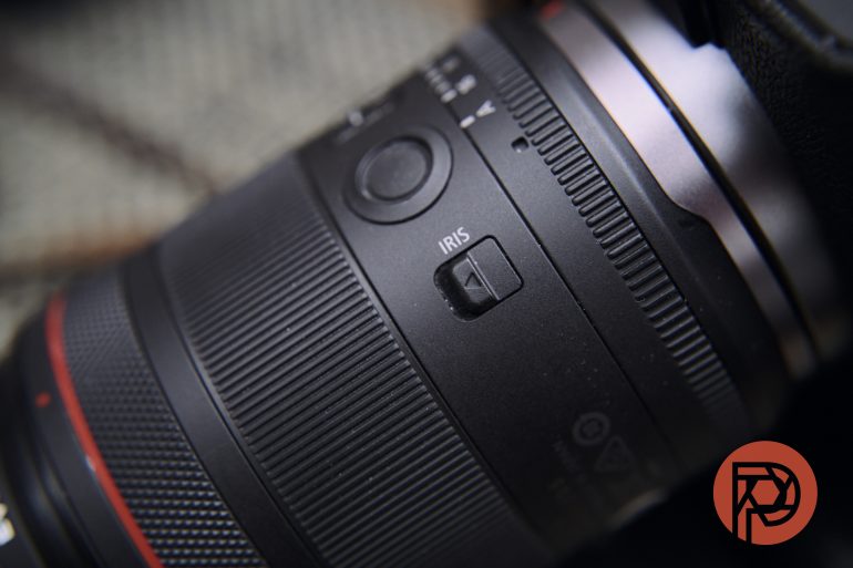 Close-up of a camera lens showing a switch labeled IRIS; red logo in the bottom right corner.