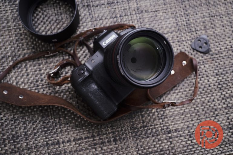 A Canon camera with a leather strap rests on a woven surface next to a lens hood and a small black item.
